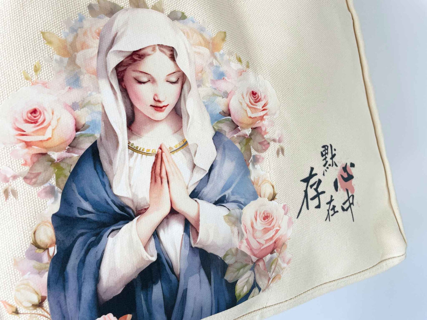 米色帆布托特包的正面下方特寫。左邊印有聖母瑪利亞祈禱的水彩圖案,她的兩旁被玫瑰花包圍,右邊印有「默存在心中」這五個字。