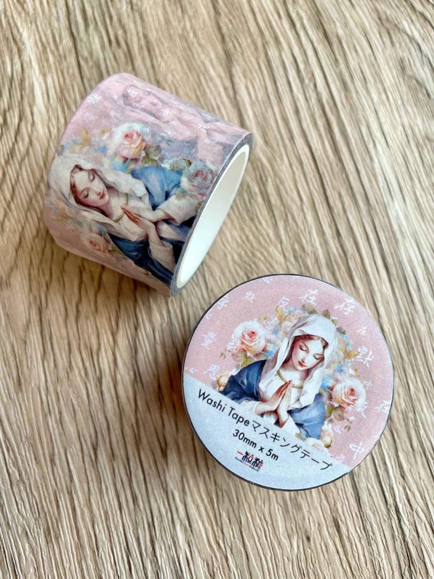 木桌上有兩卷和紙膠帶,右下方的一卷上面貼有彩紅色的圓形貼紙,裡面印有聖母瑪利亞的祈禱圖案,背後印有「默存在心中,反覆思量」的白色文字。在貼紙下方印有「Washi Tapeマスキングテープ 30mm x 5m 一粒糖」的字樣。左上方的一卷紙膠帶被豎立在桌面上,可以看到這款紙膠帶上的聖母瑪利亞祈禱圖案和粉紅色的背景。很美喔!