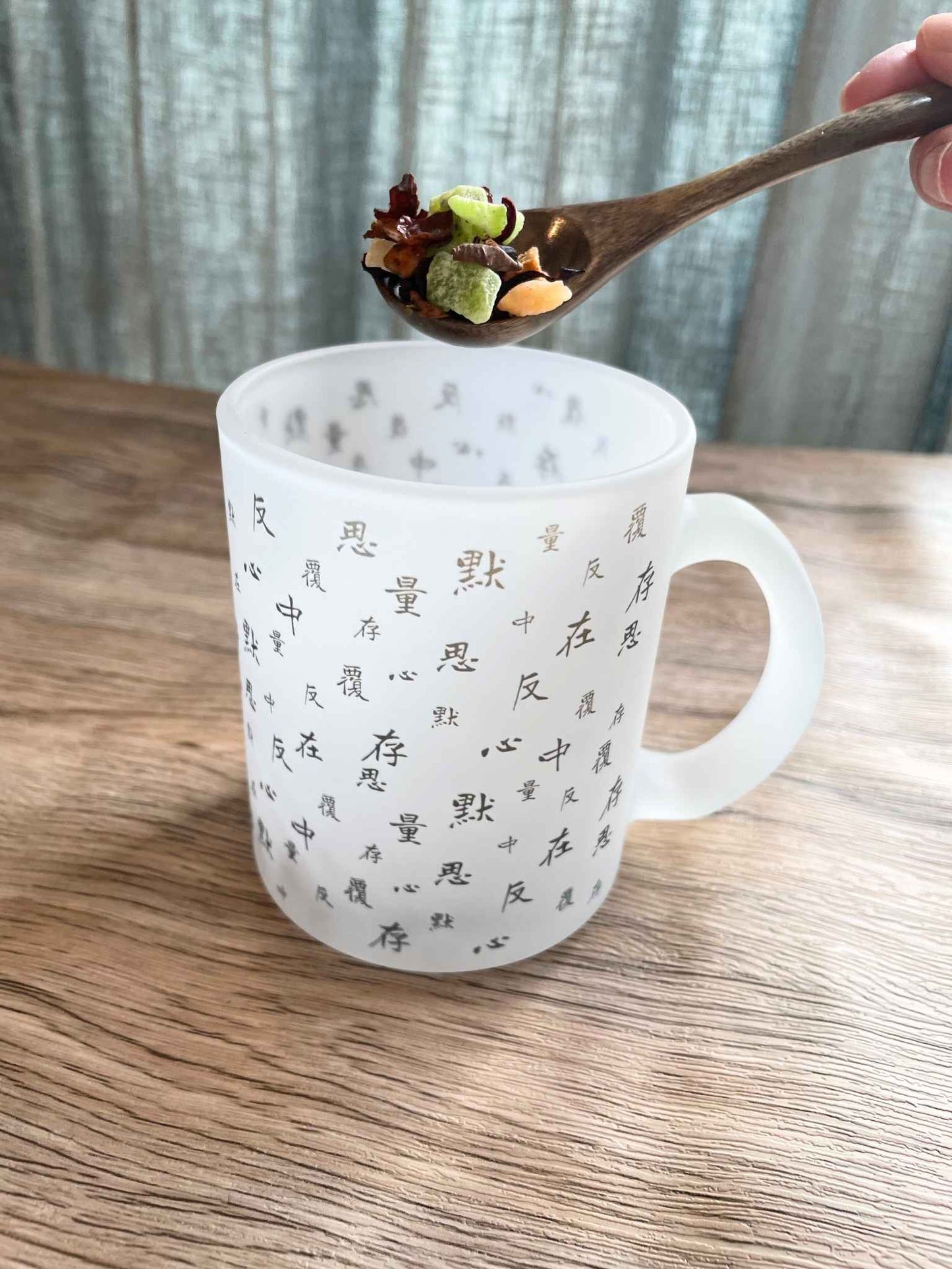 木桌面上有一隻白色磨砂馬克杯,杯子上面印有不斷重復的「默、存、在、心、中、反、覆、思、量」的藏青色文字。背景是綿麻灰藍色的窗簾。一隻手握着一支胡桃木顏色的小茶匙,正準備倒入不同顏色的花果茶乾料。