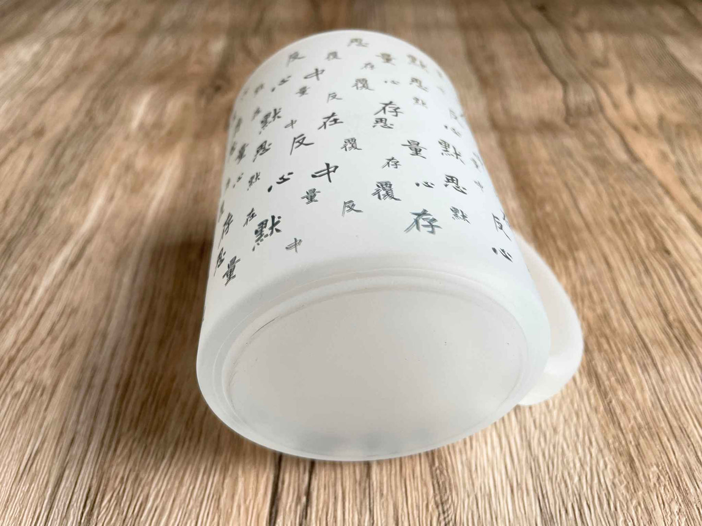 木桌面上有一隻白色磨砂馬克杯躺平在上面,杯底向下。杯子上面印有不斷重復的「默、存、在、心、中、反、覆、思、量」的藏青色文字。