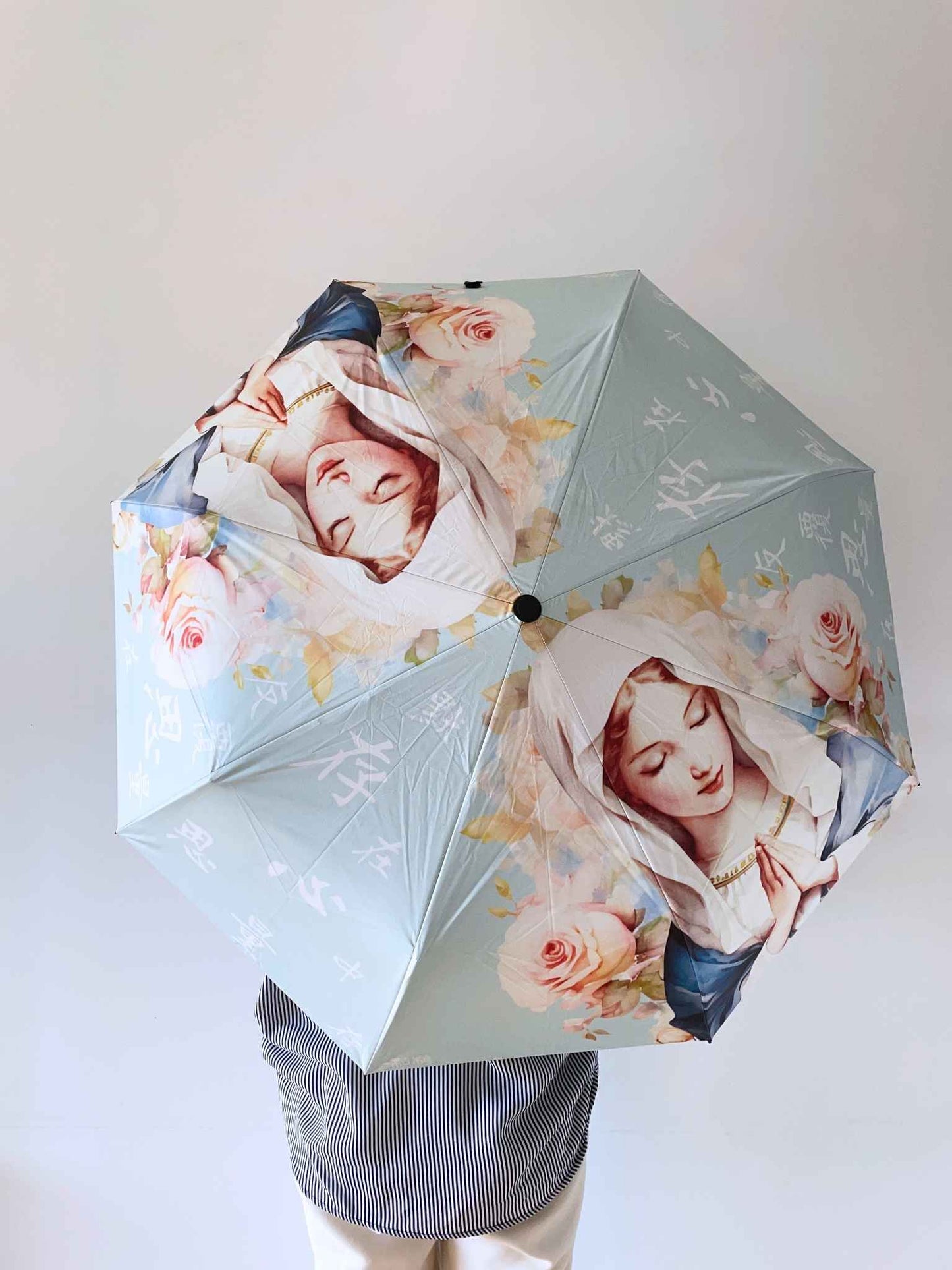 一個身穿深藍色直紋白襯衫的女生,撐着一把雨傘,雨傘面整面朝向背面,面向前面的一面白色牆。這把傘滿版印有聖母瑪利亞祈禱的水彩圖案,她的兩旁被玫瑰花包圍,上面印有「默存在心中,反覆思量」的文字,底色是淡Tiffany Blue。