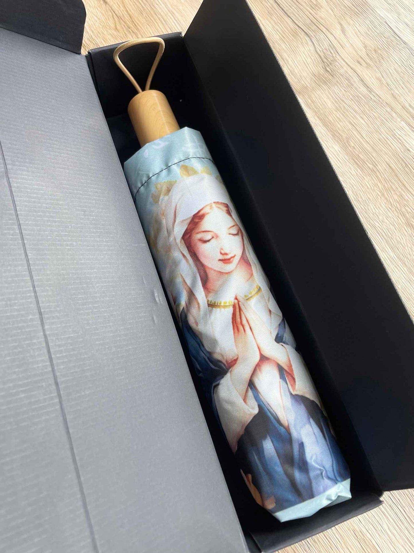 在木桌上一個深藍色紙盒被打開,裡面有一把未打開的摺疊雨傘。雨傘手把是木色的,上面有一個同色的橡筋圈。雨傘套上印有聖母瑪利亞祈禱的粉色圖案。