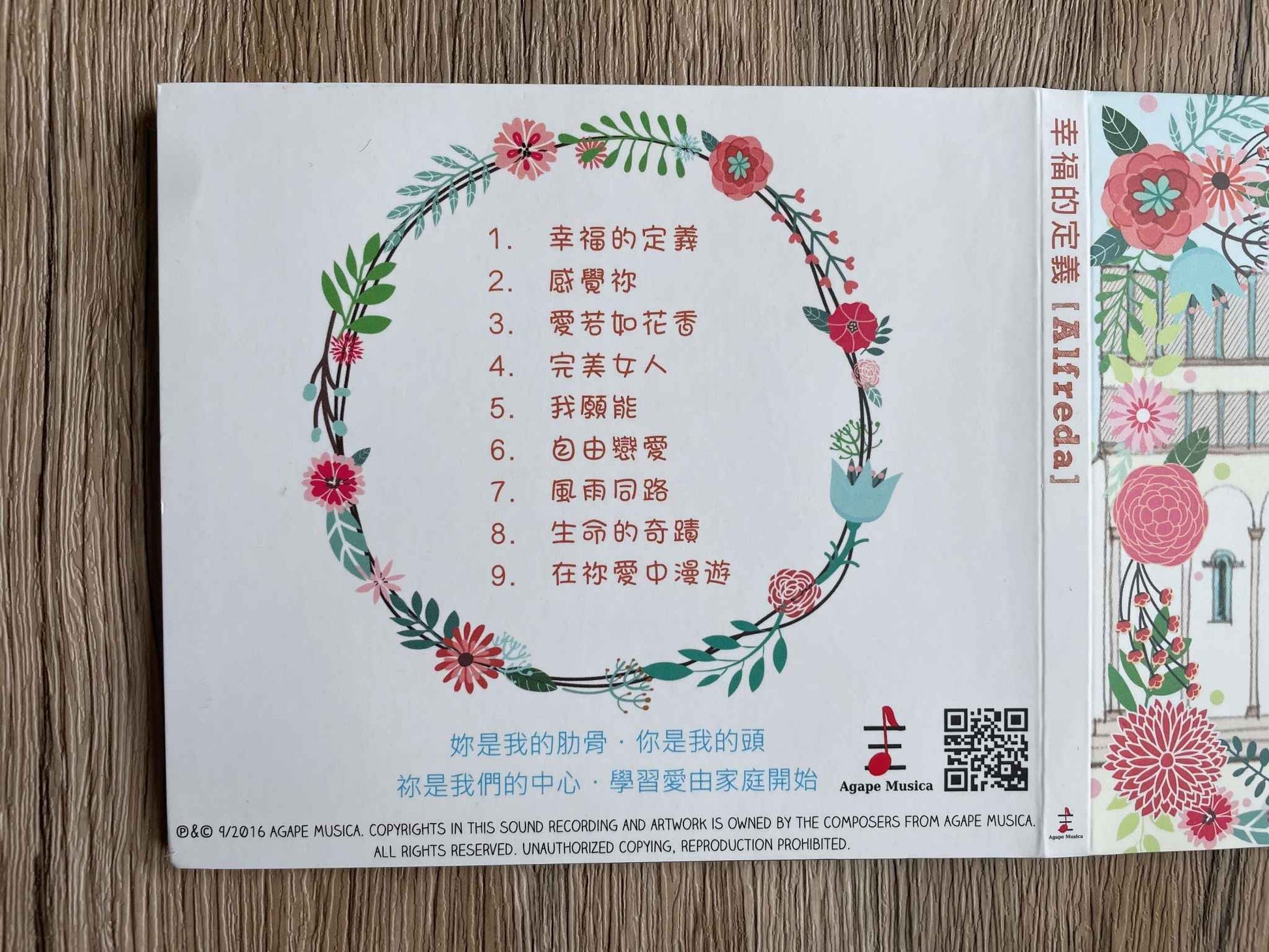 「幸福的定義」CD包裝的白色背面，中間有粉色花朵的花圈，裡面有九首歌曲的歌名。右下方是Agape  Musica的logo及QR Code。下方印有粉藍色字句:妳是我的肋骨，你是我的頭，祢是我們的中心，學習愛由家庭開始。